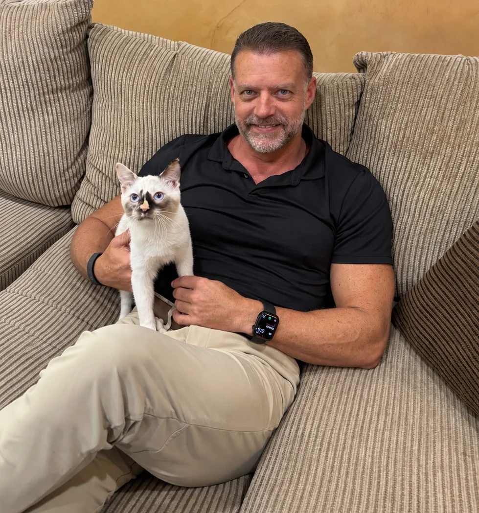 Dr. Steve Pelton sitting on a couch, holding a young cream and seal point kitten with blue eyes.